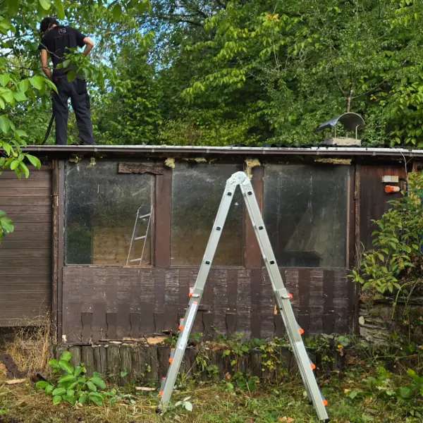 Altes Gartenhaus aus Holz mit trüben Fenstern, eine Person steht auf dem Dach