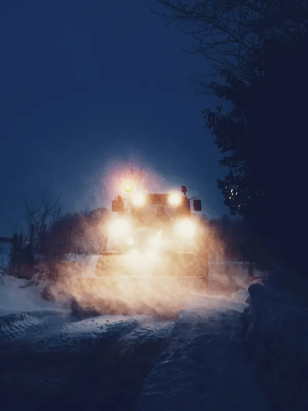 Räumfahrzeug mit eingeschalteten Scheinwerfern auf einem verschneiten Weg vor Sonnenaufgang