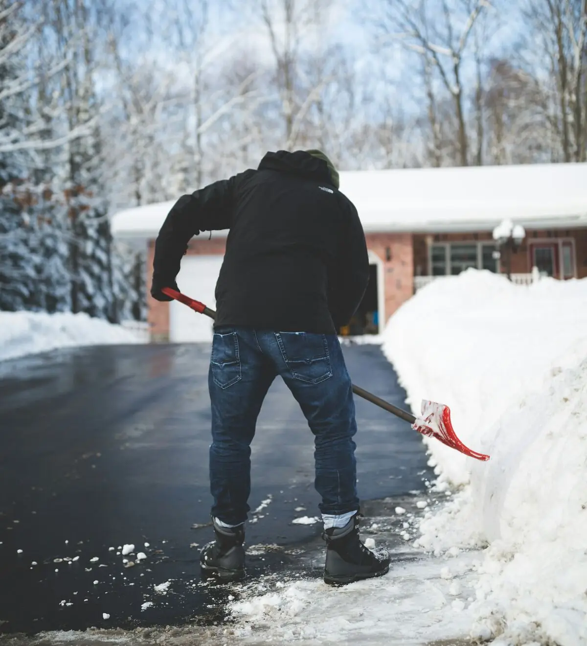 Person schiebt mit einem roten Schneeschieber eine Einfahrt frei, Rückenansicht